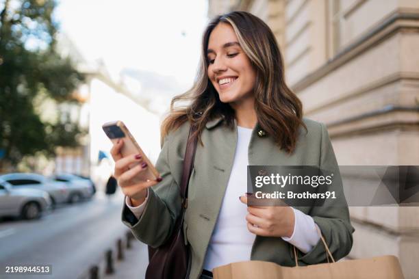 stylish young woman using smartphone and holding shopping bag in city - cliente imagens e fotografias de stock