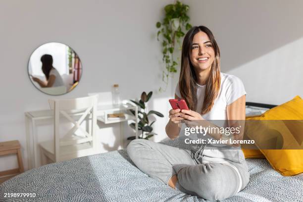 young woman relaxing on bed using smartphone in modern bedroom - rapariga no quarto tecnologia imagens e fotografias de stock