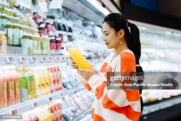 mature asian woman grocery shopping in supermarket. she is reading the nutrition label on the bottle of fresh orange juice. - sugar free stock pictures, royalty-free photos & images
