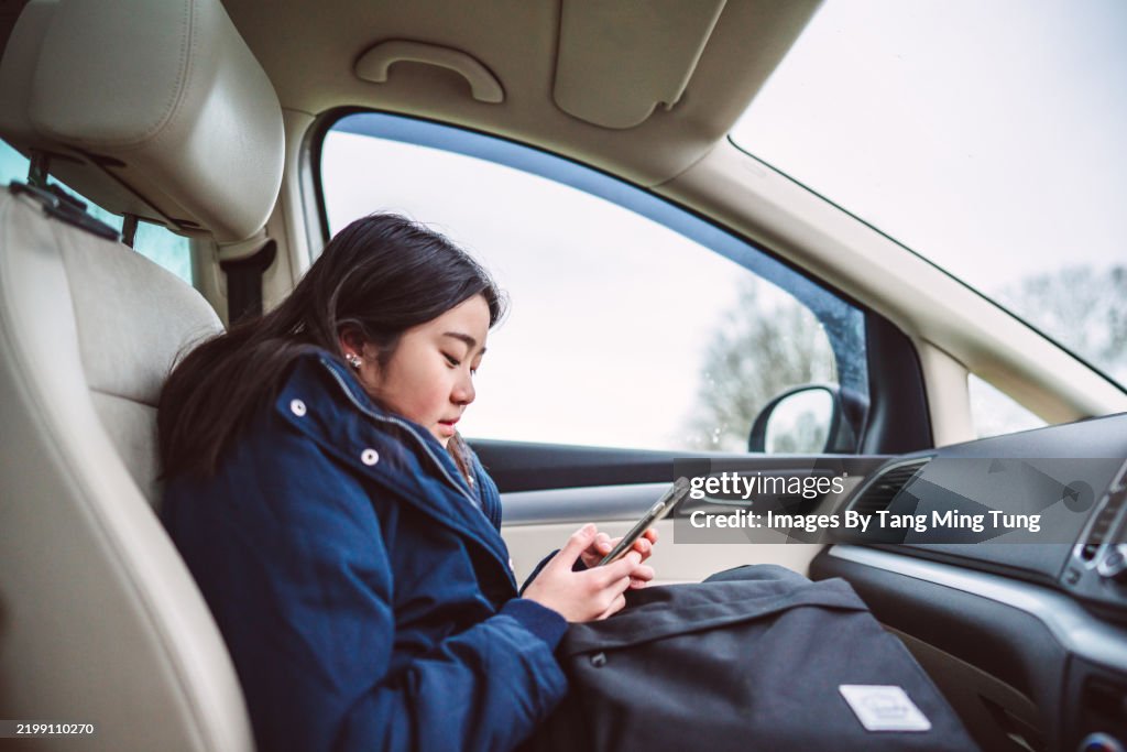 Cheerful teenage girl in school uniform using her smart phone in car in the morning