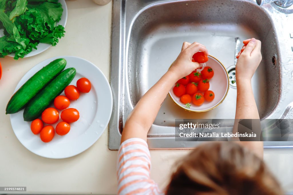Happy little chef washes vegetables before making a fresh salad, enjoying the cooking process. Hands close up and top view