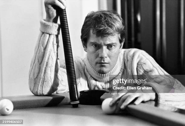 View of American actor Troy Donahue , a pool cue in one hand and a cue ball on the other, as he crouching down at the corner of a pool table, Beverly...