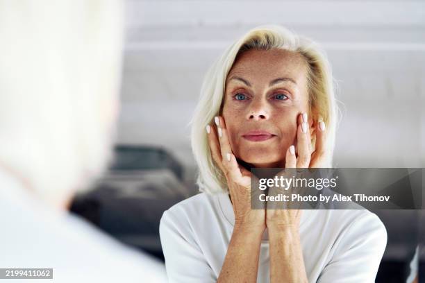 older woman standing in front of mirror, gently touching her face with hands, close up - crema viso foto e immagini stock