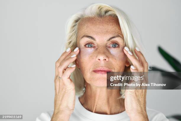 older woman looking at camera and applying on her face hydrogel under-eye recovery patches - proceso de envejecimiento fotografías e imágenes de stock