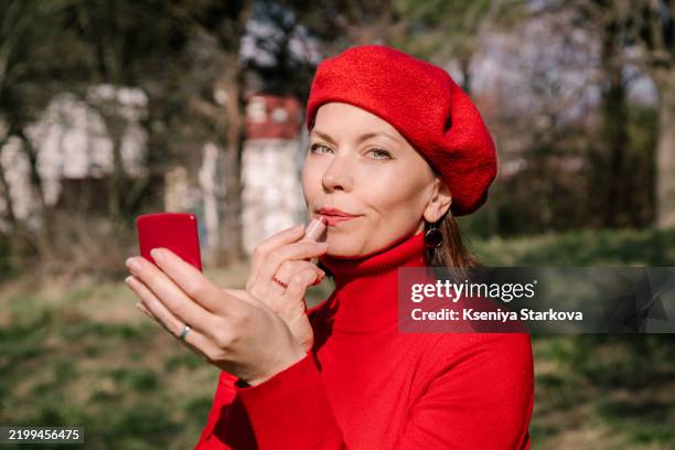 beautiful middle-aged european woman with short hair in a red beret and a red sweater paints her lips with lipstick in the park looking in a red mirror - beret stock pictures, royalty-free photos & images