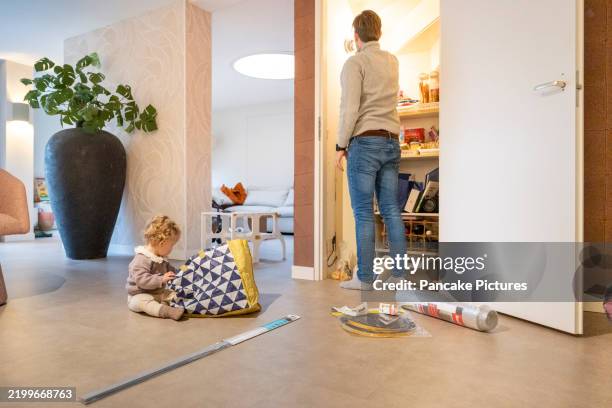 child playing with a bag beside an adult in a modern living room with open pantry. netherlands - food pantry bags stock pictures, royalty-free photos & images