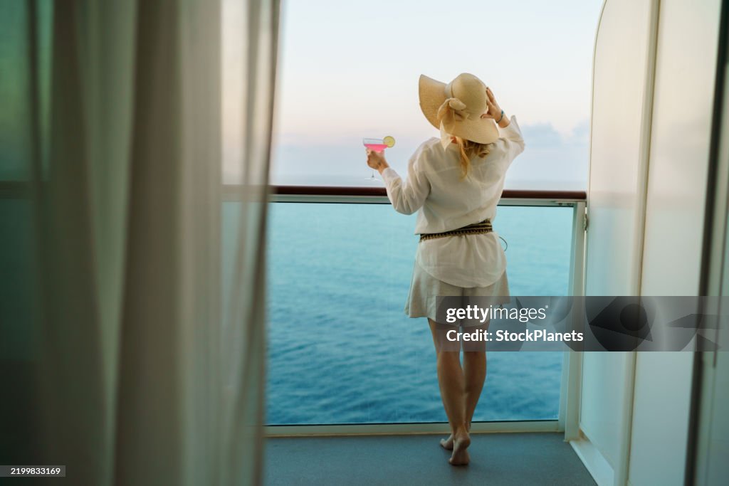 Frau mit Sonnenhut und Cocktail auf dem Balkon eines Kreuzfahrtschiffes