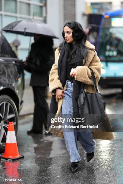 Guest wears a brown fluffy coat, a black top, a black large bag, blue denim pants / jeans, ear muffs, outside Brandon Maxwell, during New York...