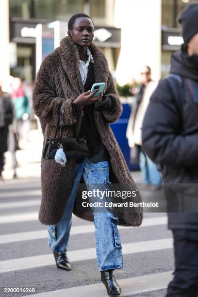 Model wears a brown faux fur coat, a crocodile pattern leather bag, blue denim pants / jeans with side pockets, leather shoes, outside Calvin Klein,...