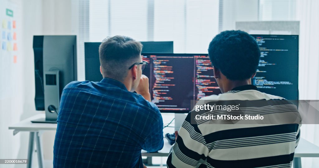 Two male software developers meeting on multiple computer screens displaying programming code, collaborating on project in office. Artificial intelligence and programming.