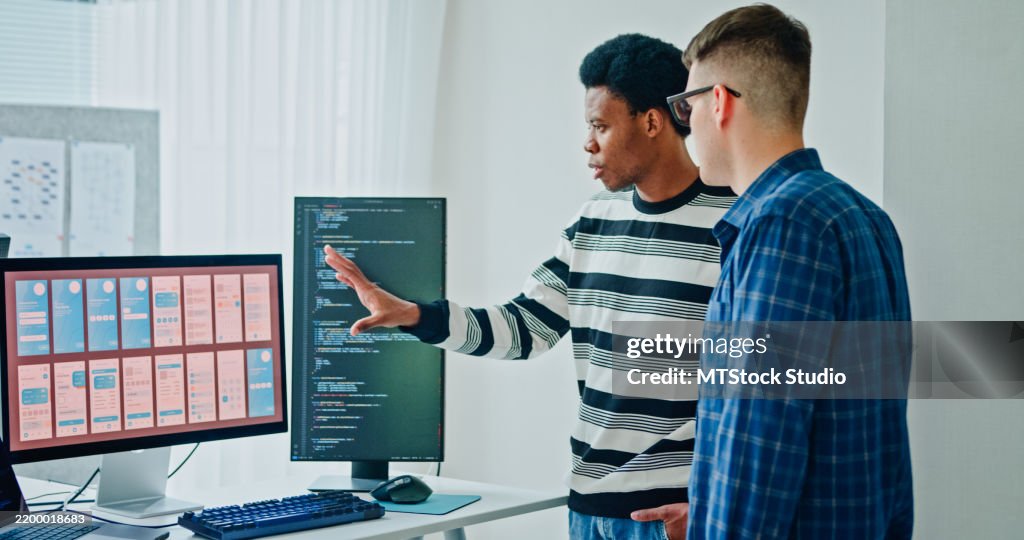Two male software developers meeting on multiple computer screens displaying programming code, collaborating on project standing in office. Artificial intelligence and programming.