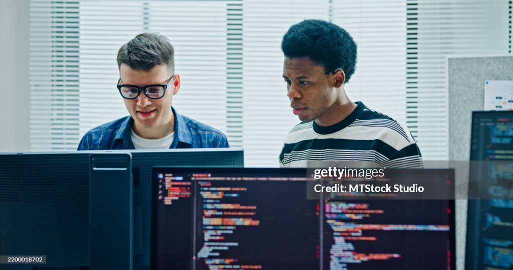 Two male software developers meeting on multiple computer screens displaying programming code, collaborating on project standing in office. Artificial intelligence and programming.