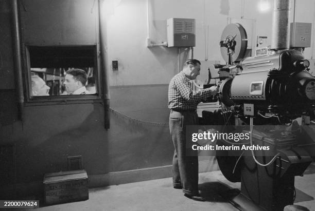 Inside a movie theater projection booth, a man adjusts a movie projector, February 9, 1958. A projected film is visible through a window on the left...