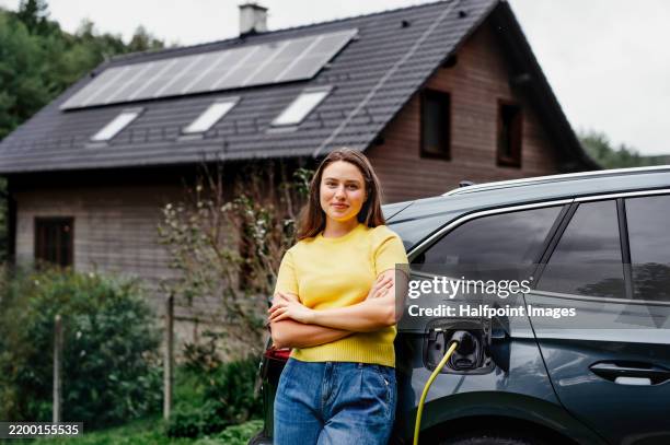 young woman is charging her electric car, parked in front of a house with solar panels on the roof. - battery storage stock pictures, royalty-free photos & images