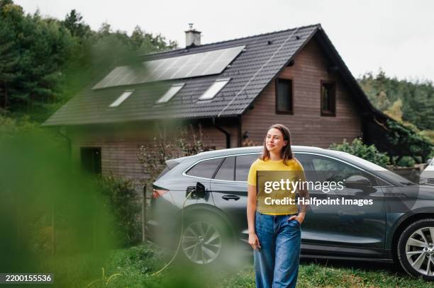 young woman is charging her electric car, parked in front of a house with solar panels on the roof. - battery life stock pictures, royalty-free photos & images