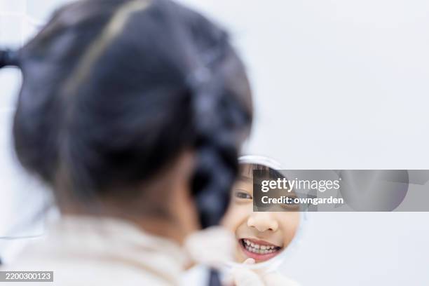 an asian small girl smiling and holding a mirror, checking her pearly white teeth at dentistry - kinderzahnheilkunde stock-fotos und bilder