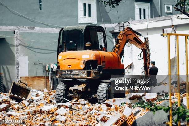 an orange excavator stands among piles of broken bricks in front of a grey building with white windows. construction site is filled with dust and debris as the machine works on demolition. - dismantling stock pictures, royalty-free photos & images