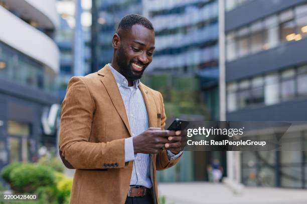smiling businessman using smartphone in modern city - brown suit stock pictures, royalty-free photos & images