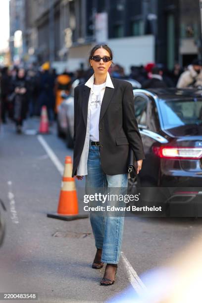 Guest wears sunglasses, a white shirt, a black oversize blazer jacket, a Calvin Klein t-shirt, a belt, blue denim pants / jeans, outside Calvin...