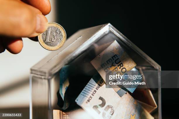 putting a coin in a piggy bank looking like a little house at home. - wohngebäudeversicherung stock-fotos und bilder