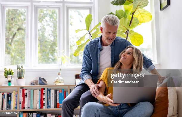 happy couple using their laptop at home and smiling - 50 54 years stock pictures, royalty-free photos & images