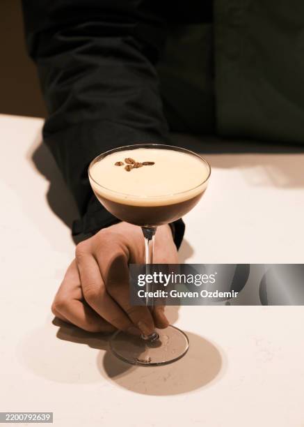 hand holding an elegant espresso martini in a coupe glass with creamy froth and coffee bean garnish, refined cocktail presentation in a stylish bar setting, luxury mixology and fine drinking concept - martini imagens e fotografias de stock