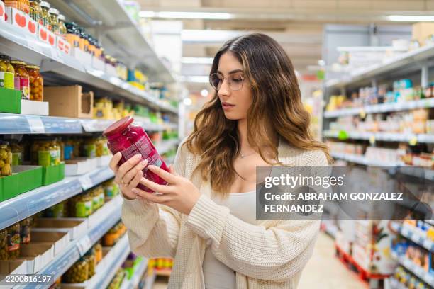 female professional studying product label, wearing eyeglasses in grocery store aisle, surrounded by packaged foods, comparing nutritional details - etiqueta nutricional imagens e fotografias de stock