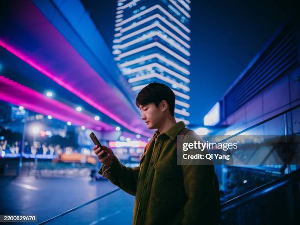 confident asian man using smartphone while standing under illuminated pedestrian bridge. in the background are tall buildings and a busy financial district - cyberpunk stock pictures, royalty-free photos & images
