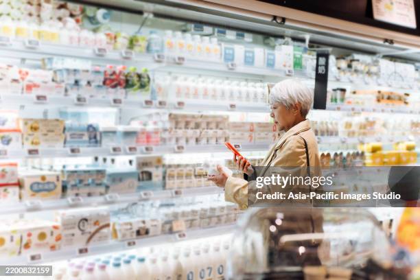 élégante femme âgée asiatique choisissant du lait frais et utilisant un téléphone intelligent pour vérifier l’étiquette nutritionnelle sur le pot. un mode de vie familial sain. faire des choix alimentaires plus sains. concept de sensibilisation d - produit laitier photos et images de collection