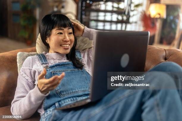 a young japanese woman is using a laptop comfortably in the living room at home. - sofa bed stock pictures, royalty-free photos & images