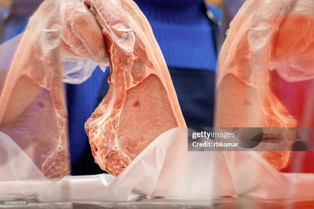 Plastic-wrapped pork chops display quality marbling at butcher counter. Fresh cuts rest on white paper with characteristic pink coloring