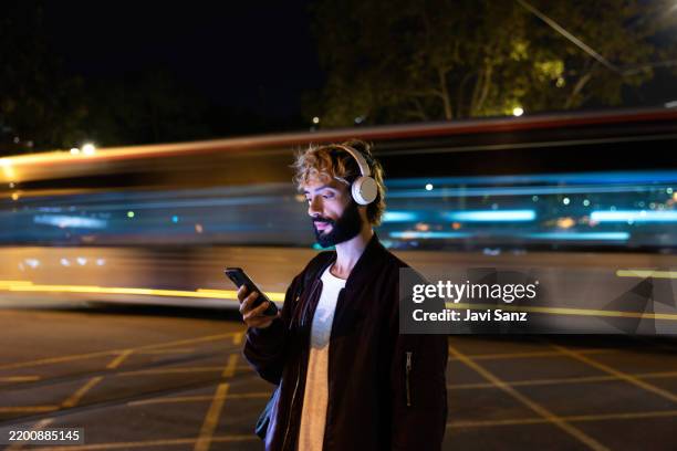 young man using smartphone with headphones and moving tram in the background at night - exposição longa imagens e fotografias de stock