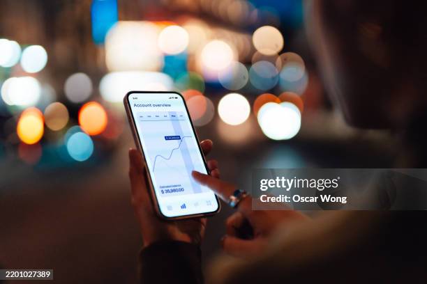 over the shoulder view of young woman managing finance and investment via trading app on smartphone - geld verdienen stock-fotos und bilder