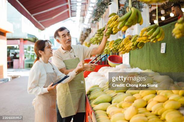 shot of two asian workers using a digital tablet while working at a fruit stall. - grocer stock pictures, royalty-free photos & images