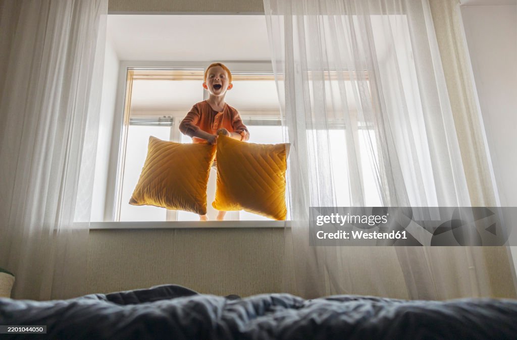 Playful boy holding pillows and standing on windowsill at home