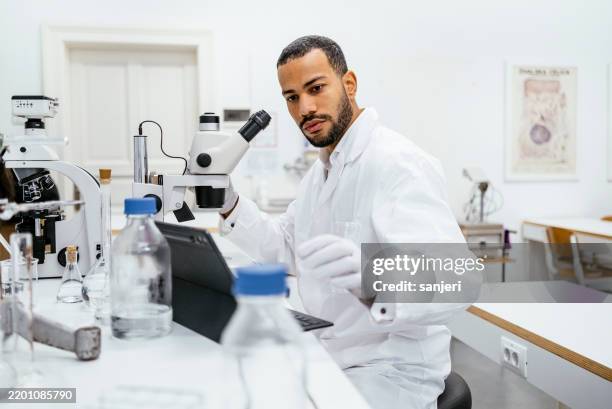 confident mixed race male student in laboratory with microscope - mikrobiologi bildbanksfoton och bilder
