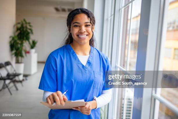 smiling female nurse in scrubs takes notes while standing in a hospital waiting room during a busy shift - enfermagem imagens e fotografias de stock