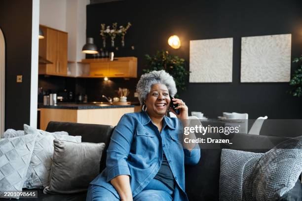 smiling mature woman with gray hair using smartphone while sitting on a cozy living room couch in a modern apartment, talking to somebody on the phone - using phone stock pictures, royalty-free photos & images