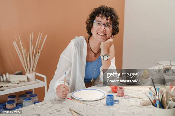 happy potter painting ceramic plate at table in workshop - renovierung themengebiet stock-fotos und bilder