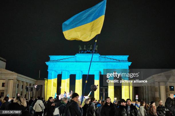 February 2025, Berlin: Numerous people stand with flags and posters on Pariser Platz, in front of the Brandenburg Gate, which is illuminated in the...