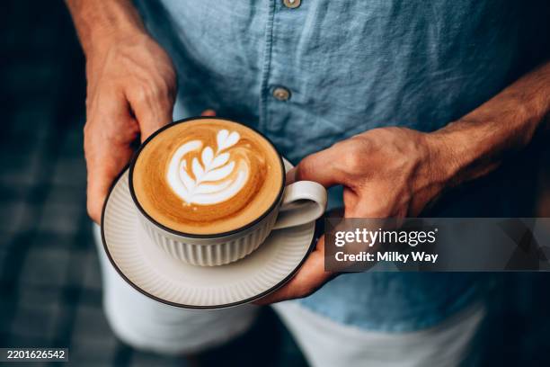 man in blue linen shirt holds a cup of cappuccino with latte art. hands gently support the saucer, emphasizing a calm and relaxing moment. background is softly blurred. - frothy drink stock pictures, royalty-free photos & images