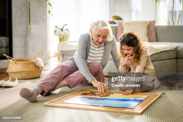 caucasian grandmother and granddaughter solving puzzle together in cozy living room - puzzle stock pictures, royalty-free photos & images
