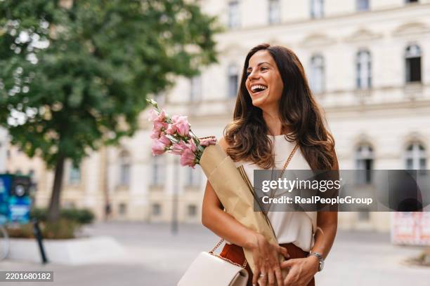 glückliche frau, die in der stadt spazieren geht und einen strauß rosa blumen trägt - blumenstrauß stock-fotos und bilder