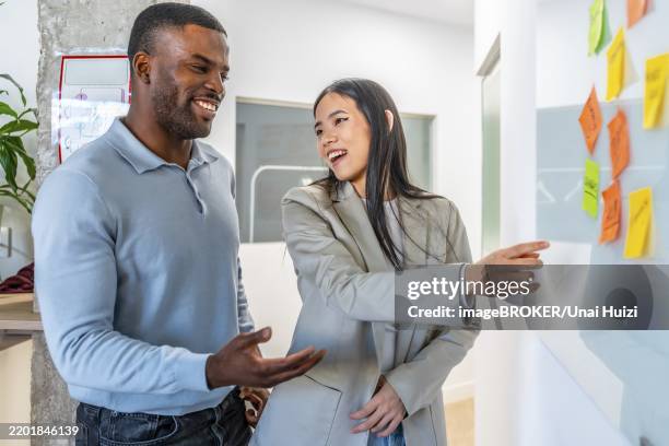 two smiling businesspeople discussing work related tasks pointing at sticky notes on whiteboard in modern coworking office - small difference stock pictures, royalty-free photos & images
