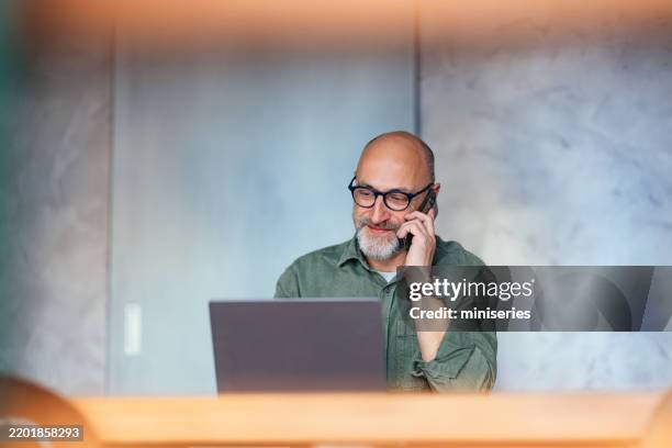 mature male professional working at laptop while talking on phone at office - reading glasses stock pictures, royalty-free photos & images