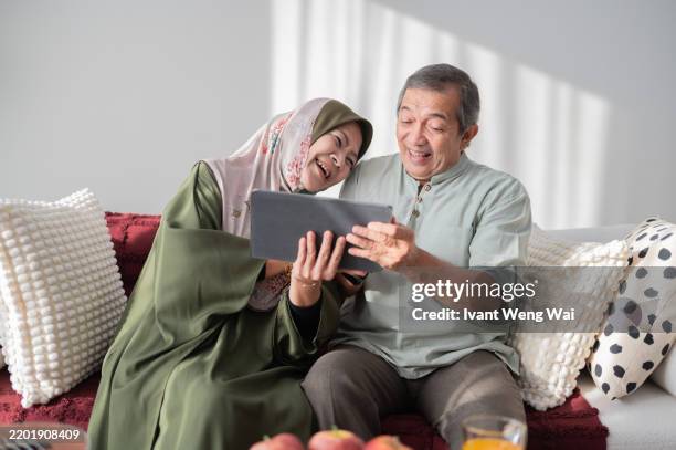 asian malay senior active couple at home sofa using smart tablet surf net sharing the moment stock photo - malay people stock pictures, royalty-free photos & images