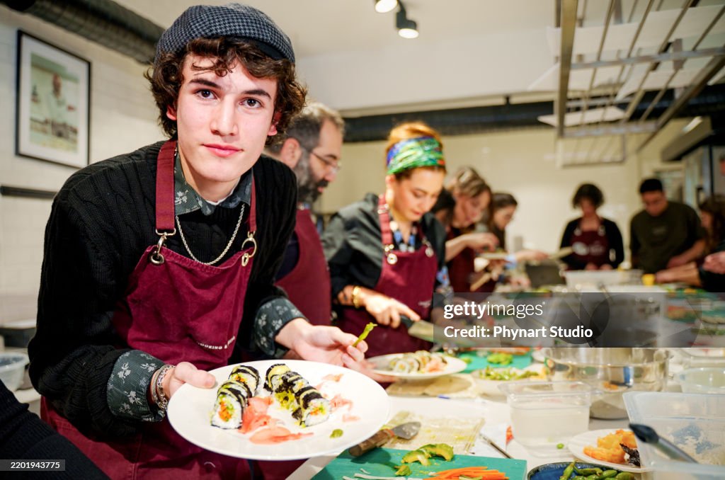 Ein junger Mann posiert mit seinem Teller, während eine bunt gemischte Gruppe von Personen mit Schürzen während eines praktischen Kochkurses sorgfältig Sushi-Rollen zubereitet.