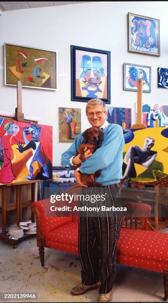 Portrait of English artist David Hockney, a dog in his arms, as he stands in front of a chaise longue in his home, Los Angeles, California, 1987.