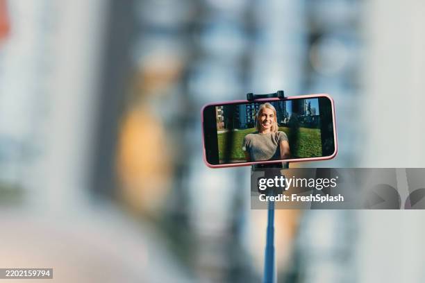 woman smiling on phone screen using a selfie stick in urban park - selfiestick stockfoto's en -beelden