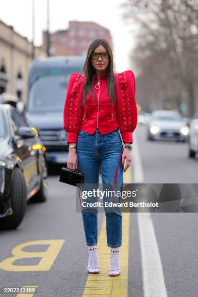 Guest wears black sunglasses, bright cherry red puffy sleeve zipped jacket, navy blue form fitting denim jean pants, gold bracelets, a black gold...